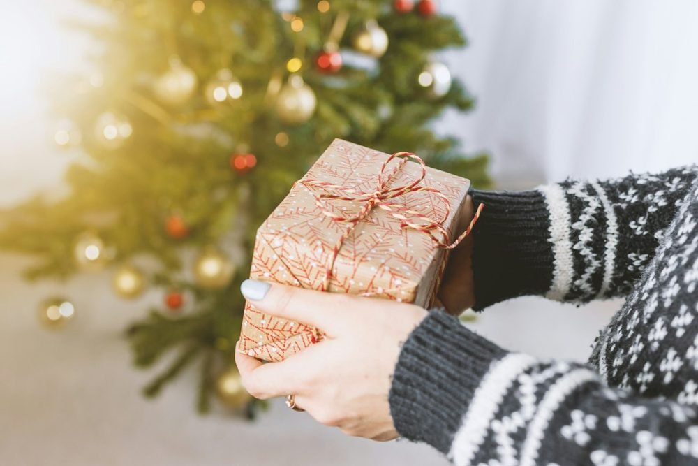 Mains qui tendent un cadeau emballé, devant un sapin de Noël
